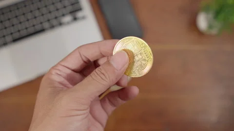 Close-up of a hand holding a gold Bitcoin coin on a work desk 库存影片 334771216