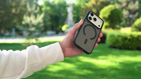 Close-up of Hand Holding Smartphone in Dark Case at Sunny Park Stock Footage 334615813