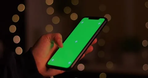 Close-up of a hand holding a smartphone with a green screen in bokeh lighting Stock Footage 339072140