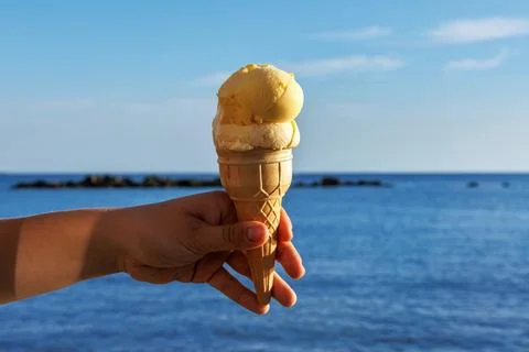 Close up of a hand holding a vanilla ice cream cone against a calm blue sea.. Stock Photos