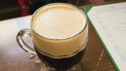 Close-up of a hand lifting a frothy dark beer mug from a wooden pub table, with Stock Footage 339237082