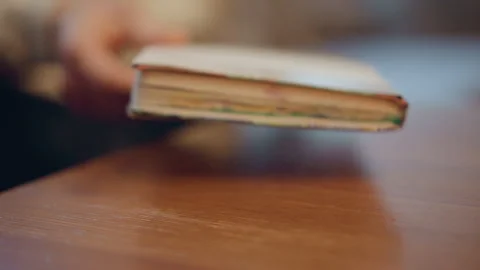 Close up of hand placing open book onto wooden table surface with soft lighting Stock Footage 347981689