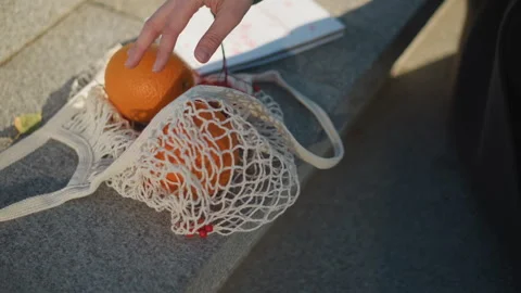 Close up of hand placing oranges into eco friendly net bag on sunlit steps with Stock Footage 346657450