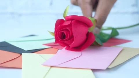 Close up of hand placing red rose flower on top of colorful envelopes pile .. Stockbeeldmateriaal 333158642