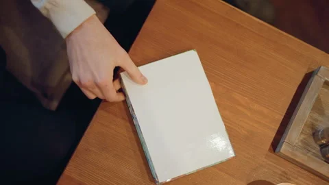Close up of hand placing white book alongside other items on wooden table with Stock Footage 346738385