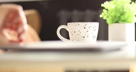 Close-up of a hand reaching for a coffee mug next to a laptop and plant Video stock 339075279