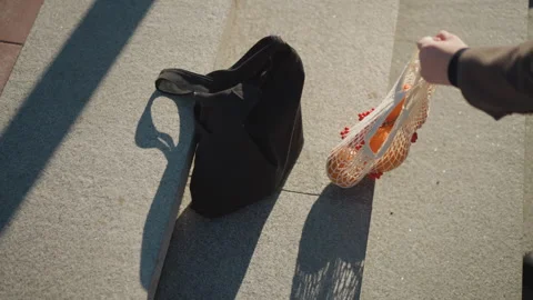 Close up of hand reaching for net bag with oranges beside black tote on concrete Vidéo 347718890