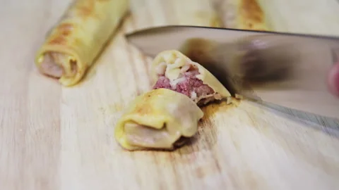 Close-up of Hand Slicing Savory Meat-Filled Crepe Rolls on a Wooden Board Stock Footage 332159722