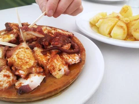 Close-up of a hand taking a piece of octopus from a portion of Galician Stock Photos
