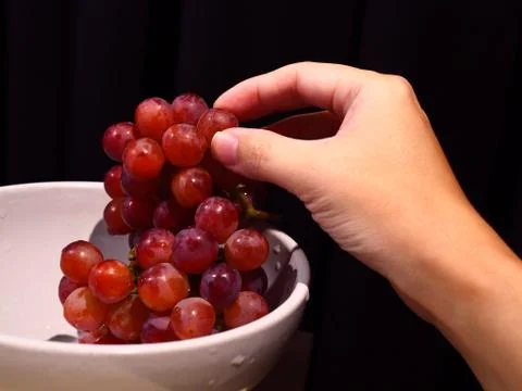 Close up hand taking red grape from ceramic bowl, black background 库存照片