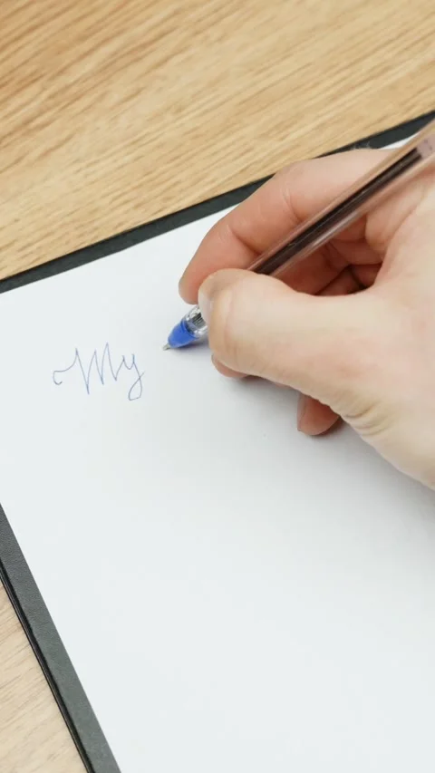 Close-up of a hand writing “My plan” with a blue pen on white paper at a wooden Stock Footage 309569216