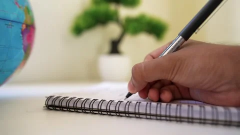 Close-up of a hand writing in a notebook with a pen, globe and plant in bac.. Stock Footage 317737116