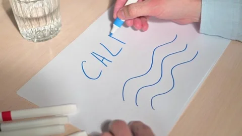 Close-up of a hand writing the word CALM with a marker next to blue waves on a Vidéo 337558640