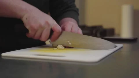 CLOSE UP OF HANDS MINCING GARLIC WITH CHEF KNIFE ON CUTTING BOARD IN KITCHEN 库存影片 336948985