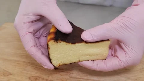 Close-up of Hands in Pink Gloves Holding a Slice of Basque Burnt Cheesecake Stock Footage 331780046