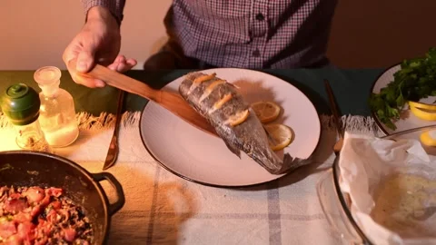 Close up of hands placing baked fish onto a plate under warm evening kitchen 库存影片 328255741