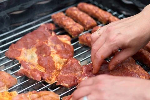Close-up of hands placing marinated pork steak and sausages on a barbecue gri Stock Photos