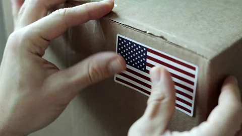 Close-up of hands placing a sticker of the USA flag on a cardboard shipping box. Stock Footage 323469316