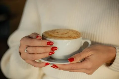 A close-up of hands with red nails holding a white cup of frothy coffee Stock Photos