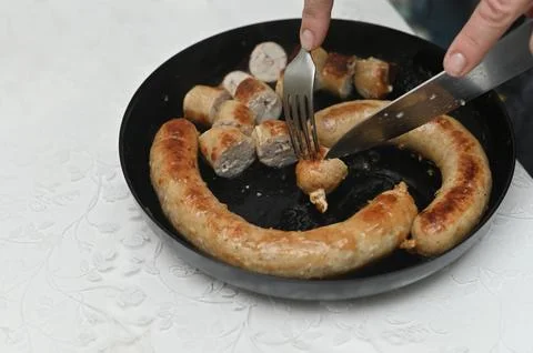 Close-up of hands slicing appetizing fried sausages with knife and fork. Stock Photos