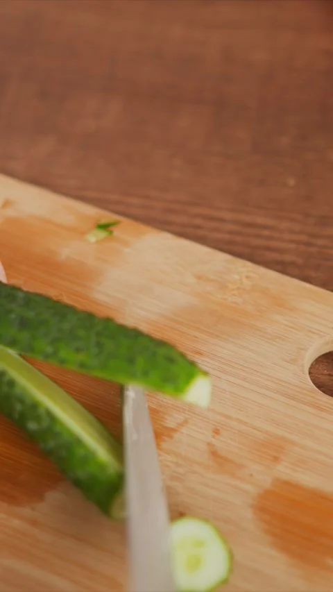Close up of hands slicing fresh cucumber on wooden cutting board with knife Stock Footage 346777128