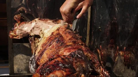 Close-up of hands slicing a piece of meat from the spit-roasted calf. Carcass of Stock Footage 248212600