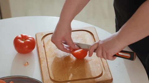 Close-up hands slicing ripe tomato on wooden cutting board in bright kitchen Stock Footage 345673315