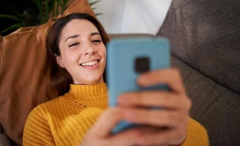 Close up hands of smiling and relaxed girl using cell phone lying on sofa in Stock Photos