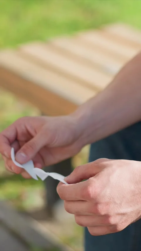 Close up of hands tearing small paper object in outdoor setting with wooden Stock Footage 345051433