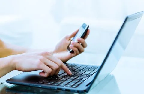 Close-up of hands typing on laptop computer while using a cell phone. Stock Photos