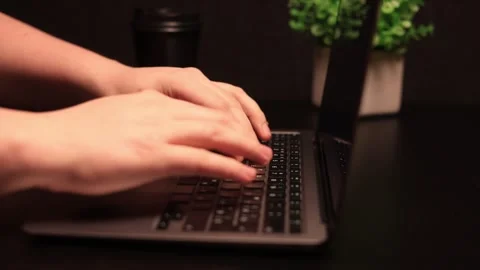 Close-up of hands typing on a laptop keyboard with a coffee cup and plant in the Stock Footage 338432517