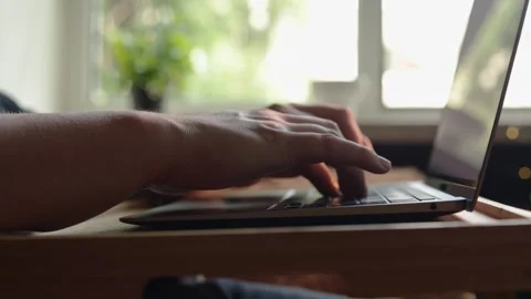 Close-up of hands typing on a laptop keyboard with a blurred window background Stock Footage 340264692