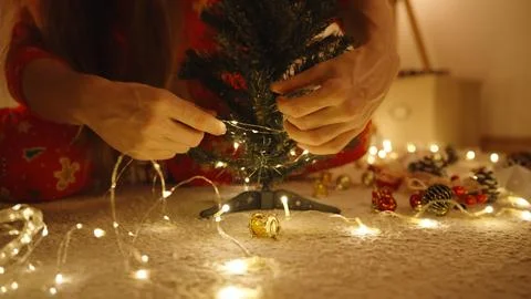 Close Up of Hands Wrapping String Lights Around Mini Christmas Tree on Carpet Stock Photos