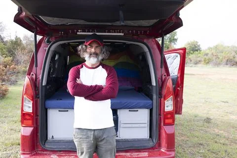 Close-up of a handsome man in a cap standing by a camping van, looking at t.. Stock Photos