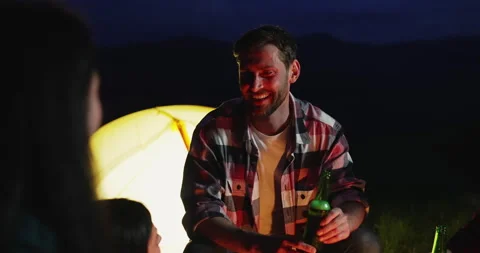 Close up of handsome young man sitting near tent at night, drinking beer and Видео 156617586