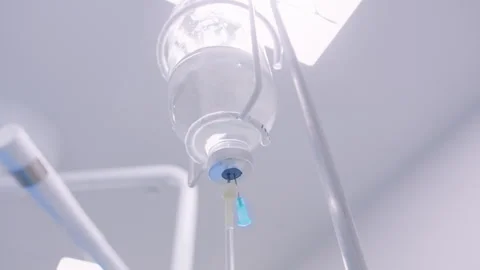 Close-Up of Hanging IV Fluid Bag with Clear Drip Chamber Under Bright Surgic 動画素材 308419862