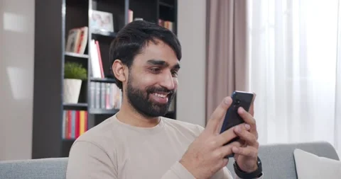 Close up of happy Arab young man sitting on sofa and chatting by messages on Stock Footage 179942504