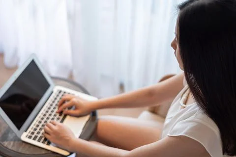 Close-up, high angle view of Asian woman sitting near curtains, using computer. Foto stock