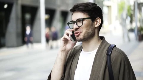 Close up of a hipster guy in glasses talking on the phone and laughing outside 動画素材 77521631