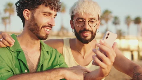 Close-up, homosexual couple using mobile phone while sitting on the beach Video stock 250840964