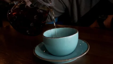 Close up human hand pouring tea into a clay blue cup from a clear glass teapot. Video stock 285126112