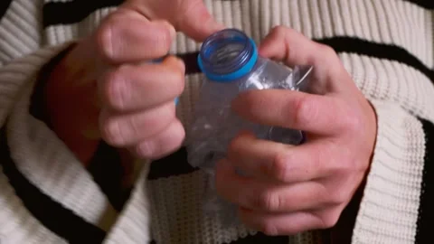 Close-up of human hands in striped sweater crushing plastic bottle Stock Footage 328237630