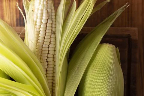 Close up image of raw white corn on the cob on wooden rustic surface. Top vie Stock Photos