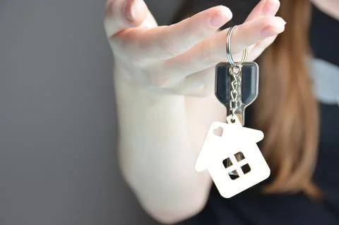 Close-up of a key with a keychain in the shape of a house in hands. Gray ba.. Stock Photos