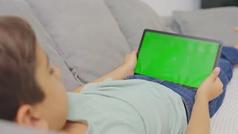 Close up kid lying on sofa, holding and tapping on a tablet with green screen. Stock Footage 255662516