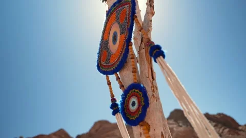 Close up knitted evil eye amulet hangs on a wooden branch stick in the desert at Stock Footage 253418786