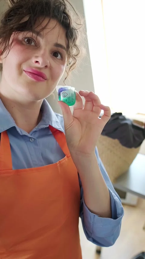 Close-up of a laundry capsule in hand, with clean folded laundry in the backgrou Stock Footage 295286497
