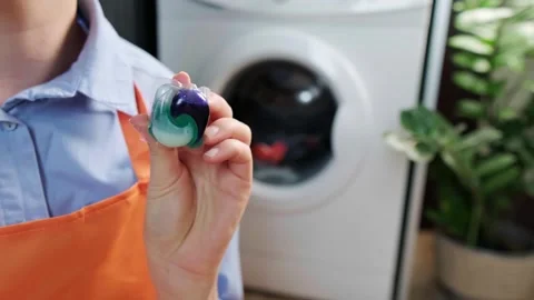 Close-up of a laundry capsule in hand, with clean folded laundry in the backgrou Stock Footage 295286942