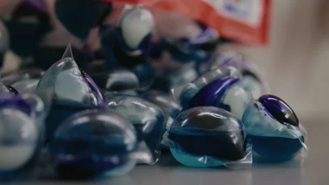 Close-up of laundry detergent pods pouring from package onto pile of more pods Stock Footage 312329011