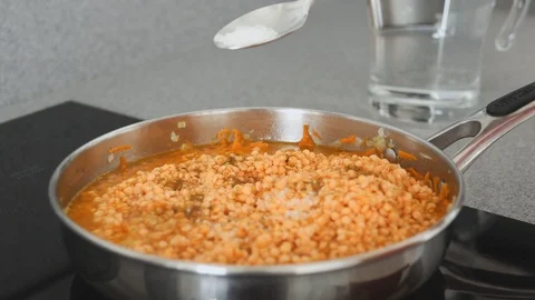 Close - up of lentils stewing in a pan. A hand with a spoon of spices appears in Stock Footage 117038906